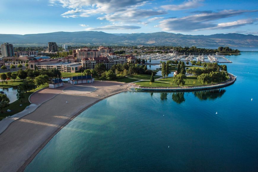 Aerial view of downtown Kelowna public waterfront with a curved beach, lakeside park and marina on Okanagan Lake