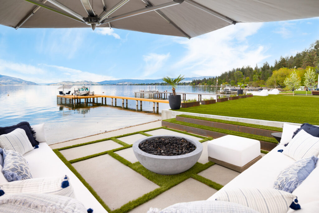 Lakefront patio with a white sectional and fire bowl beside a private dock and moored boat on Okanagan Lake