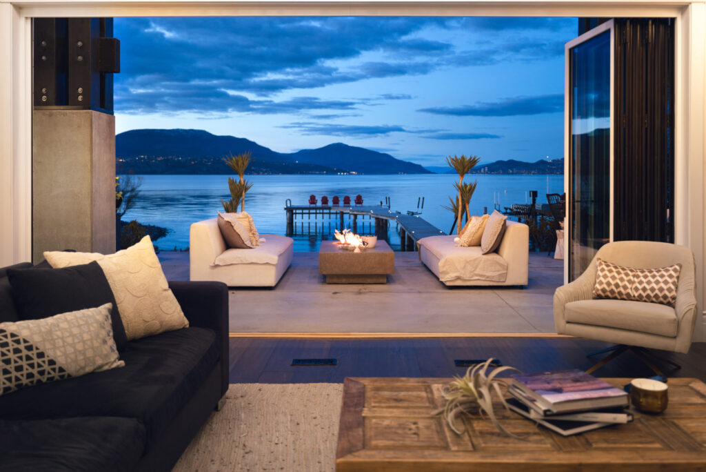 Open living room doors framing a lakeside terrace with lounge seating, a dock and Okanagan Lake at dusk