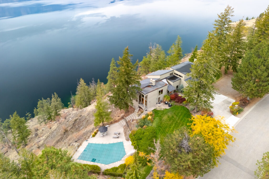 Aerial view of a high-bank estate on a treed clifftop above Okanagan Lake with a covered pool and lawn