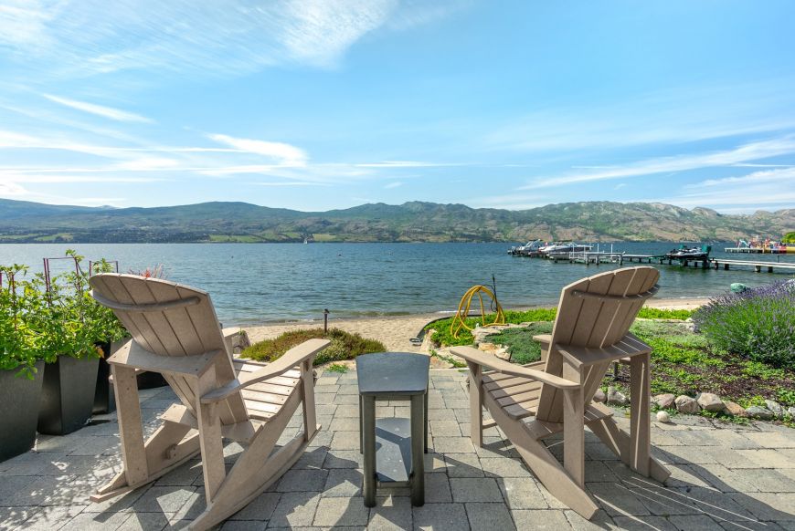 Two Adirondack rocking chairs on a stone patio facing a sandy Okanagan Lake beach, a private dock and mountains