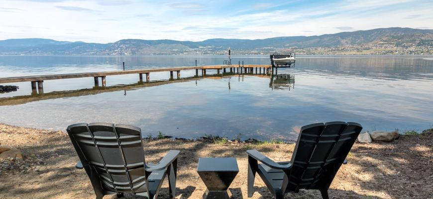 Two black Adirondack chairs on a pebble beach facing a long private dock stretching into Okanagan Lake